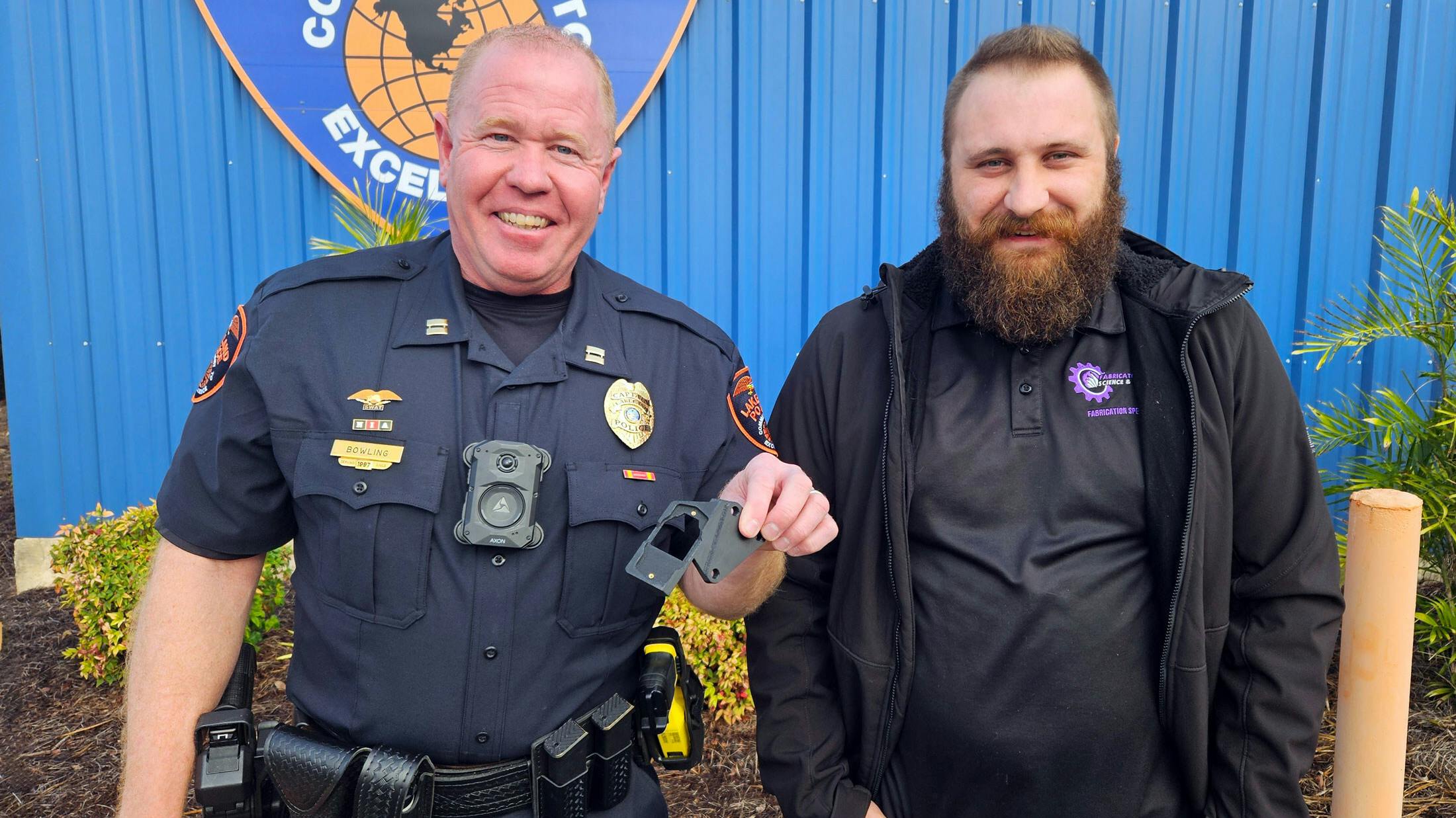 Lakeland Police Capt. Ron Bowling, Jr. (left) displays a firearm holster bracket designed and fabricated by Mike Kalman (right), mechanical engineering fabrication specialist at Florida Polytechnic University, and mechanical engineering graduate Matthew DeCicco &rsquo;24. The patent-pending design helps trigger an officer&rsquo;s body camera when it detects that a firearm has been removed from its holster.