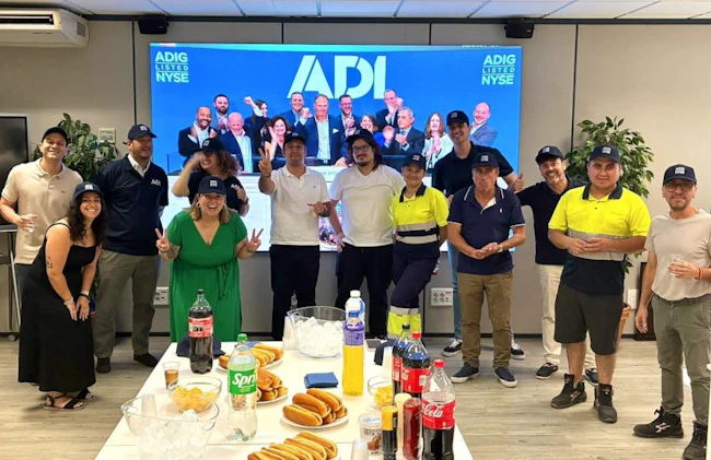 Employees at an ADI branch celebrate the company's first day of trading on the New York Stock Exchange as an independent public company, watching the exchange ceremony on a large screen.